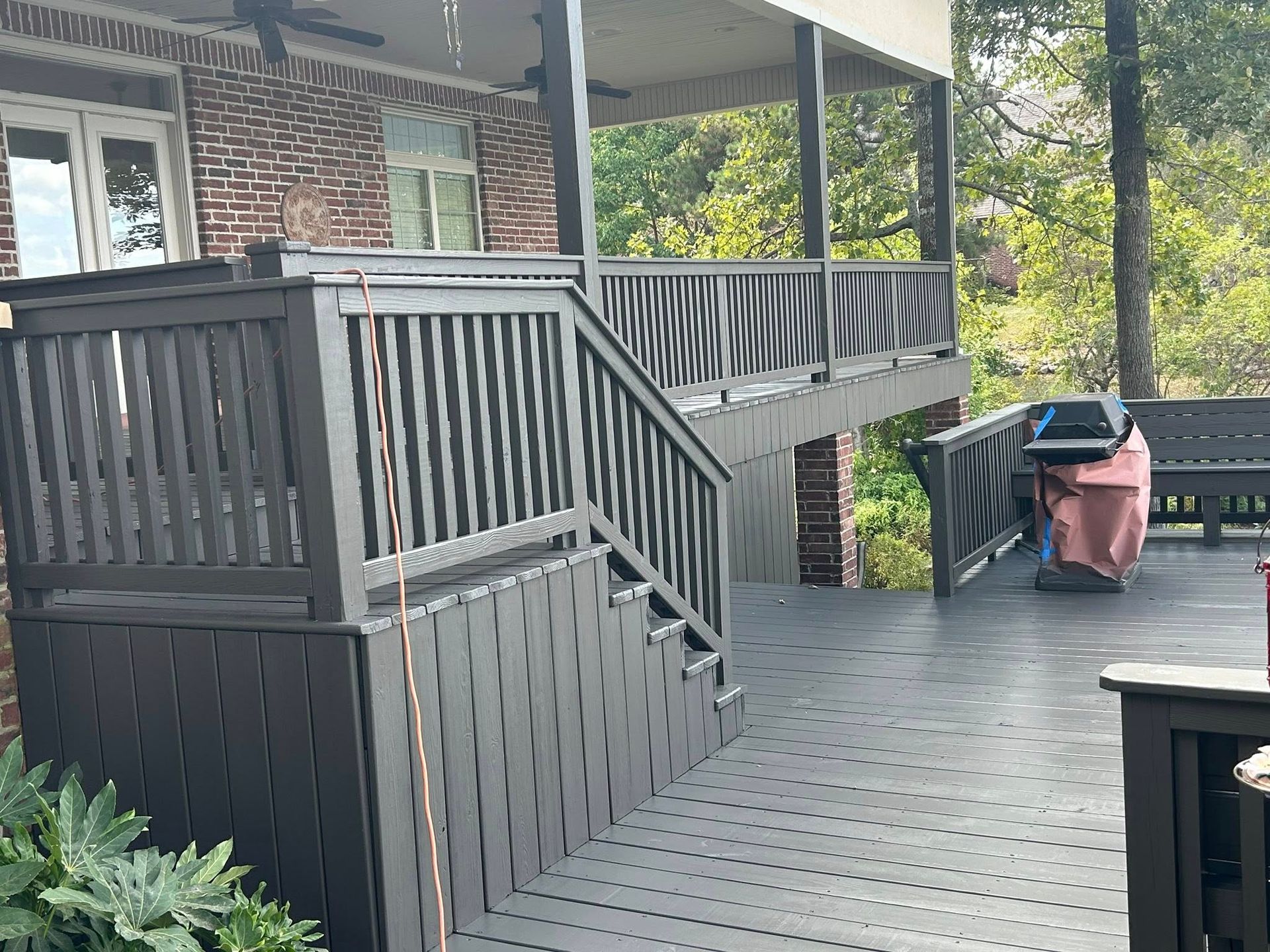 A two-level deck with gray painted railings and stairs. A brick building and trees are in the background.