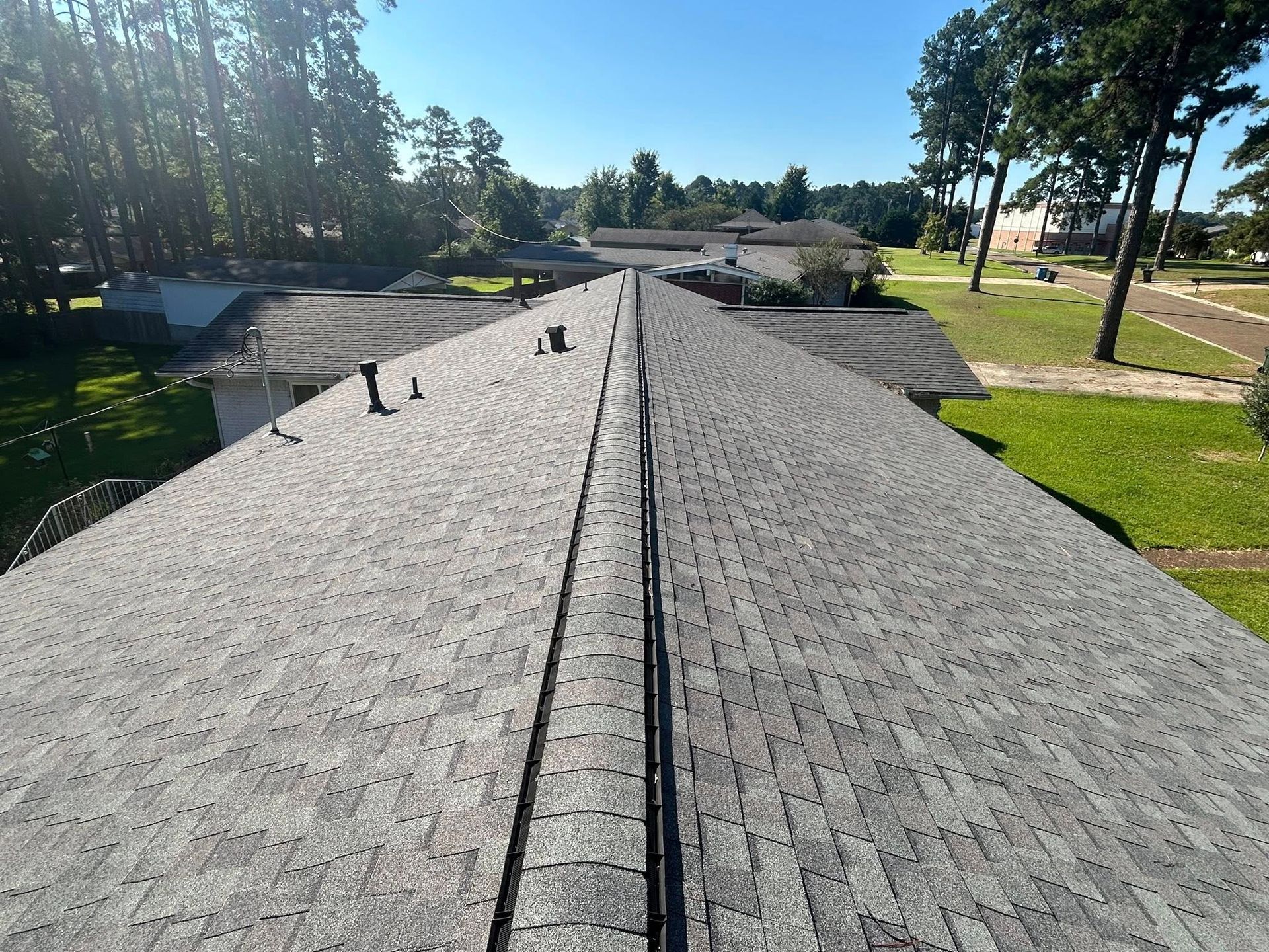 View of a gray shingled roof with a central ridge. Trees and green grass are visible in the background.