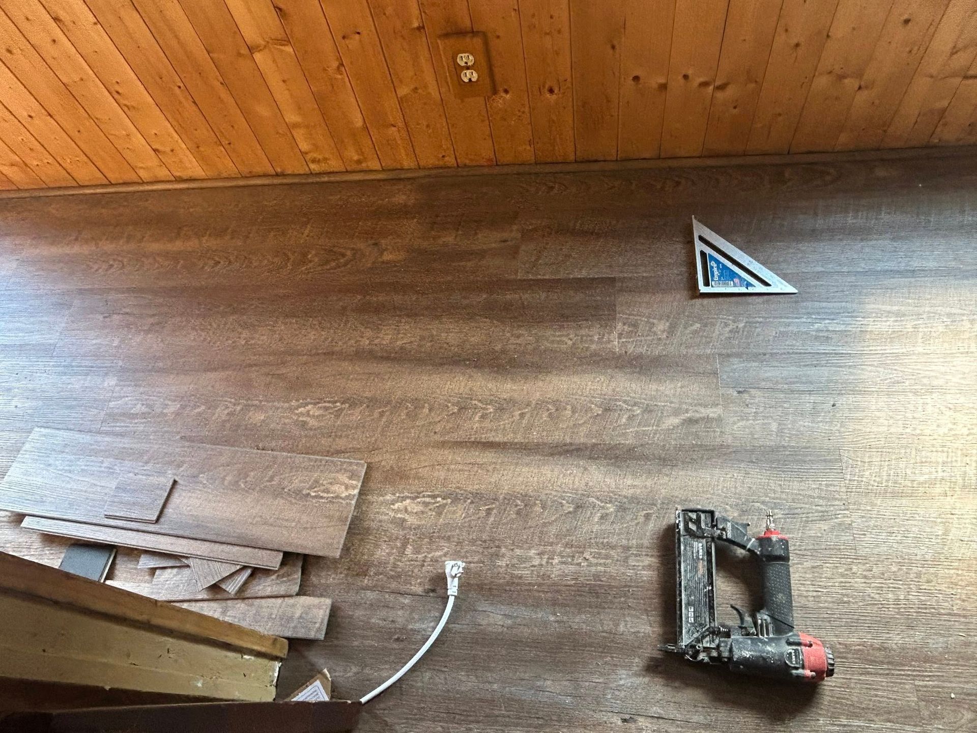 Flooring installation in progress: brown planks, nail gun, square, and light-colored wooden ceiling.