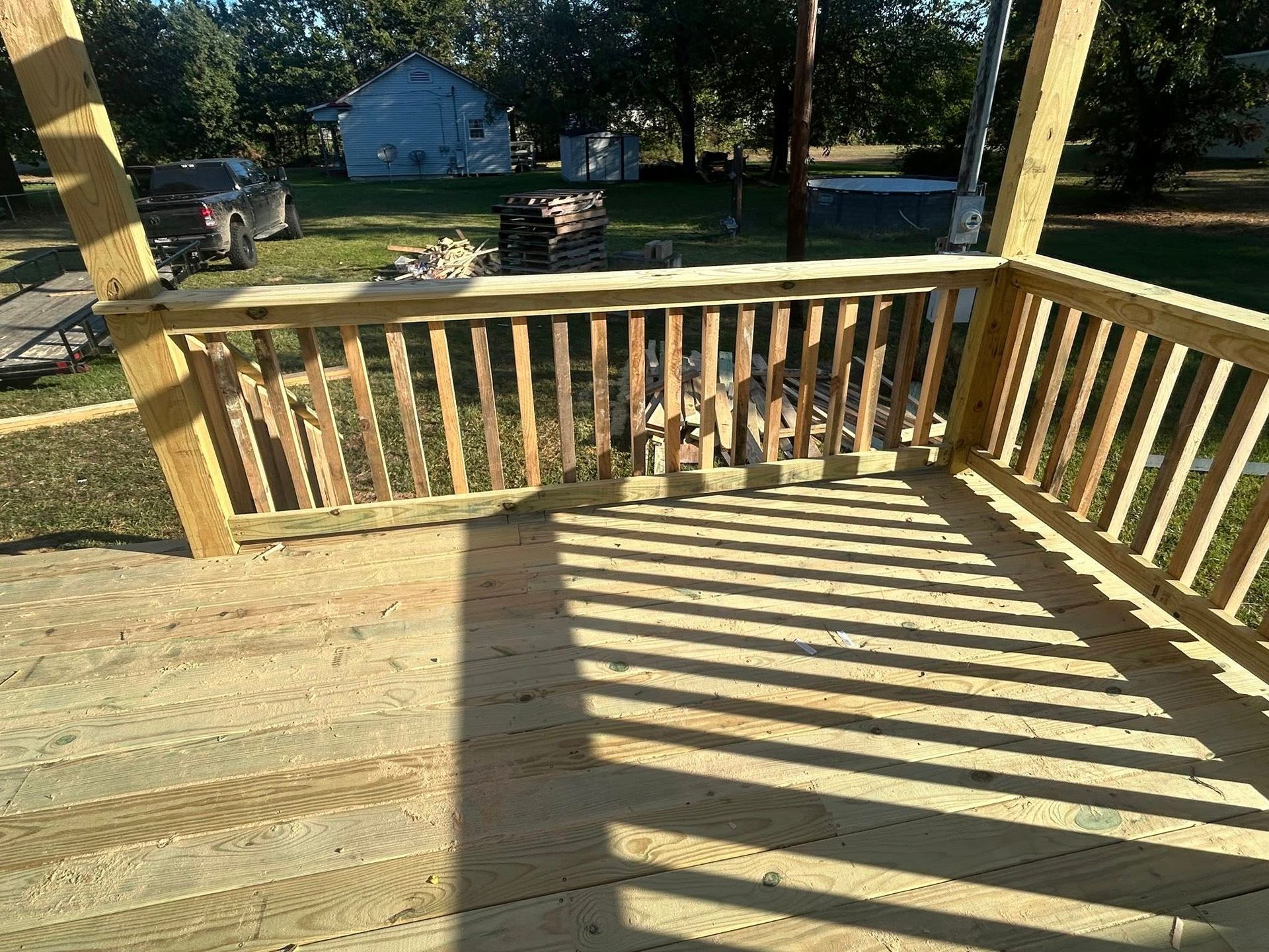 Newly built wooden deck with railings, casting shadow on the boards. Outdoor setting with houses and trees.