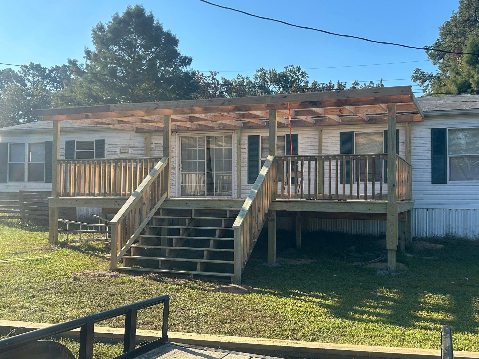 Mobile home with wooden deck, stairs, and pergola. White siding, green shutters, sunny outdoors.