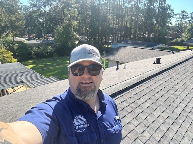 Man on a rooftop wearing a cap and sunglasses, taking a selfie. Sunlight, trees, and other roofs in the background.