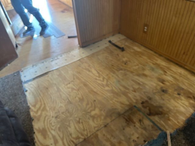 Plywood floor with some flooring removed, doorway, person’s legs in background.