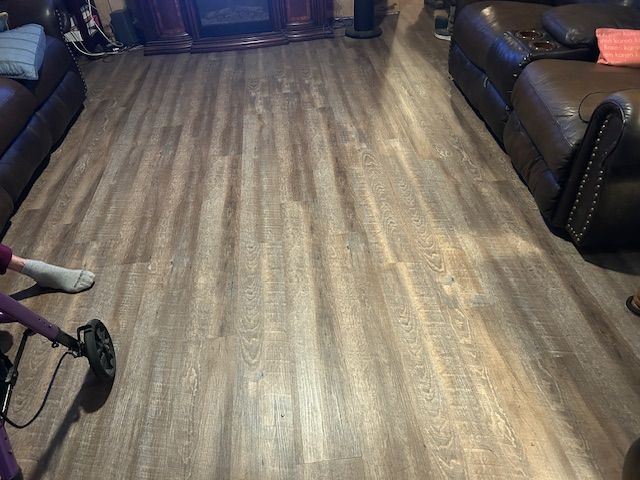 Laminate flooring in a living room with a person using a metal detector. Brown leather couches and fireplace in the background.