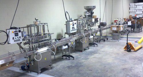 A stainless steel industrial filling and packaging assembly line in a warehouse setting.