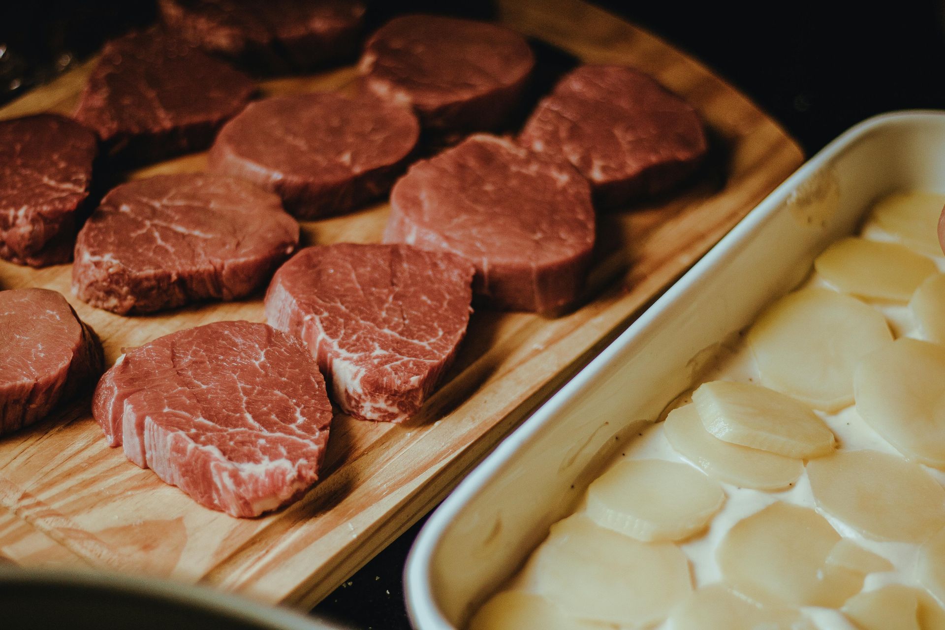 Raw steaks on a wooden board next to a casserole dish with sliced potatoes in cream sauce.