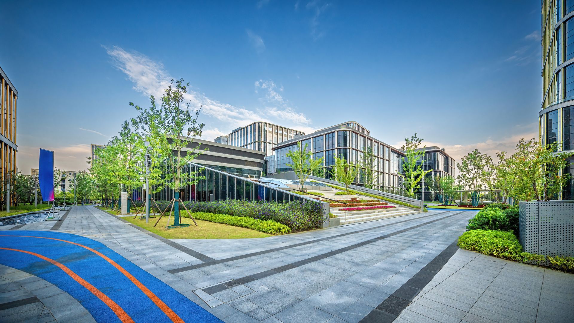 Modern building complex with glass walls, greenery, and blue sky. Brick paved walkways with a blue and orange bike lane.
