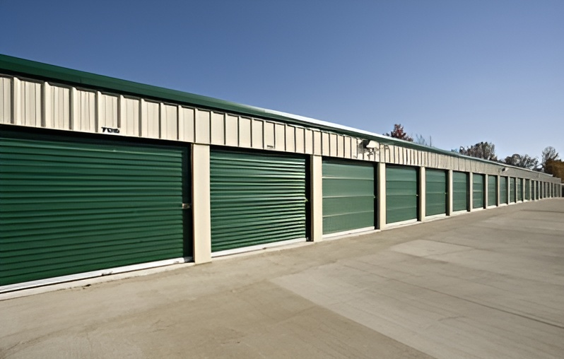 Green storage unit doors in a row under a beige and green metal roof, on a concrete pad.