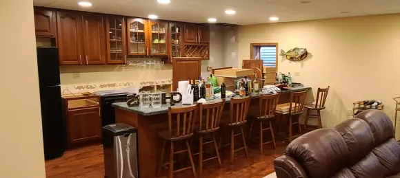 A kitchen with wooden cabinets, a bar with stools, and a leather sofa. Boxes and items sit on the bar.