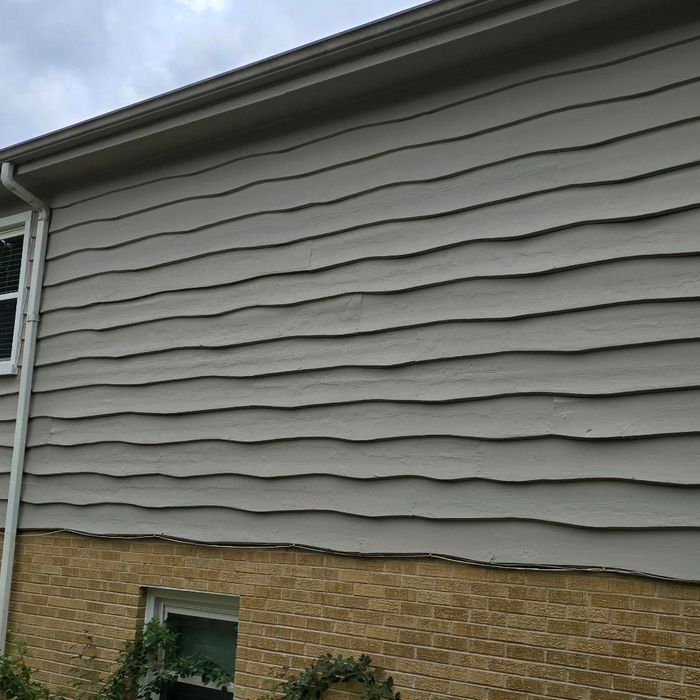 Gray wavy siding on a building above a tan brick wall; overcast sky.