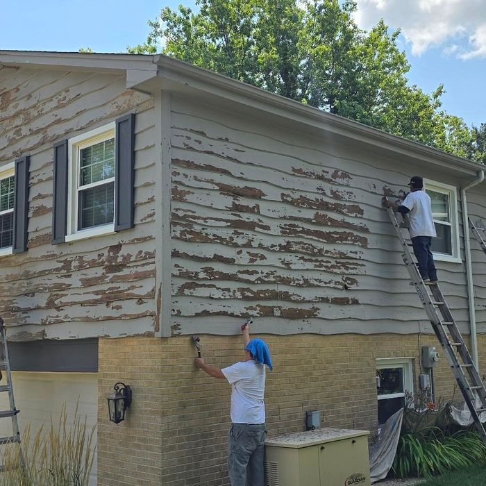 Two people paint house siding; peeling paint on a two-story home with a brick foundation.