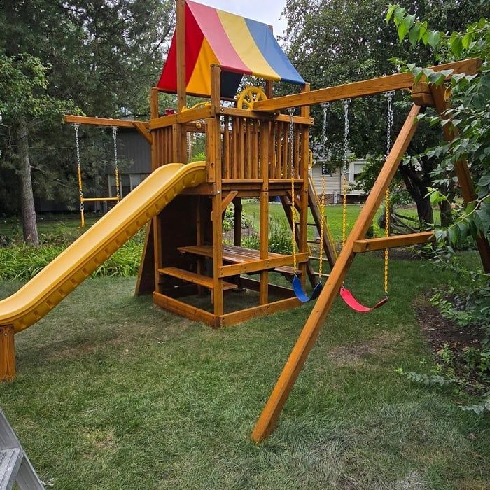 Wooden playset with slide, swings, and colorful awning in a grassy yard.