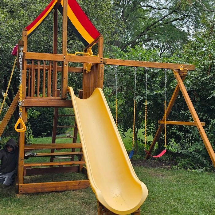 Wooden playset with yellow slide, swings, and roof. A person is kneeling next to it.
