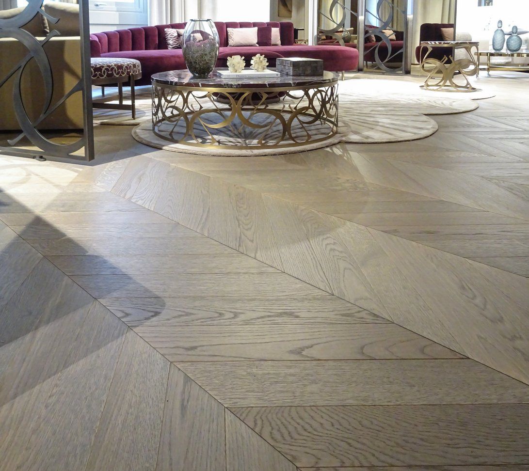 Pavimento in legno inclinato con motivo a chevron; sullo sfondo un divano morbido e un tavolino rotondo.