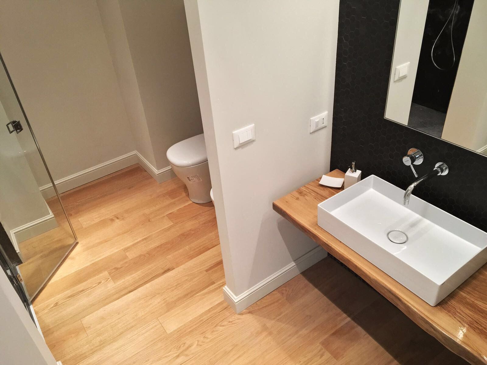 Bagno con pavimento in legno, lavandino bianco su ripiano in legno, WC sullo sfondo e doccia.