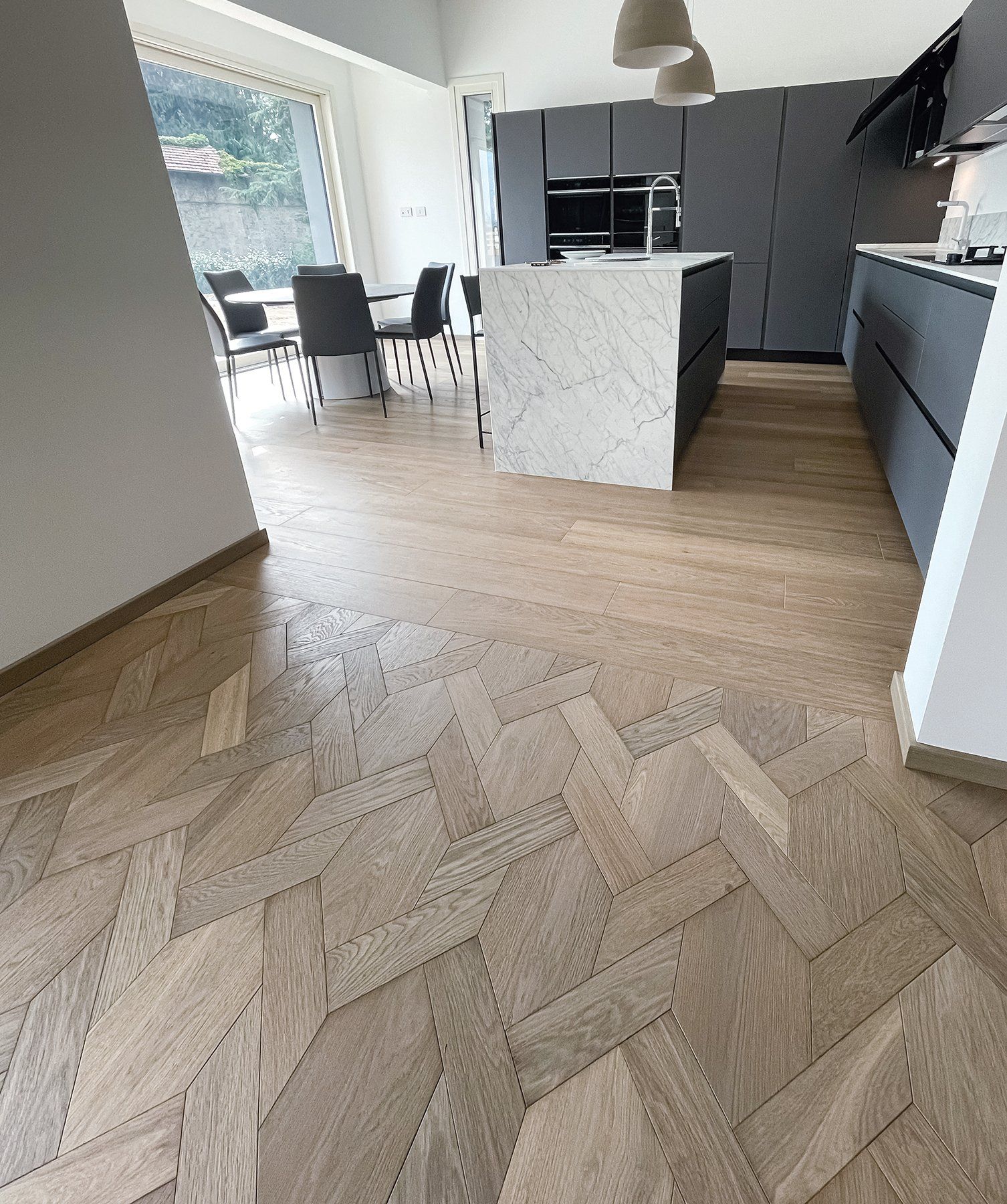 Pavimento in legno a spina di pesce in una cucina moderna con isola in marmo e mobili grigio scuro.
