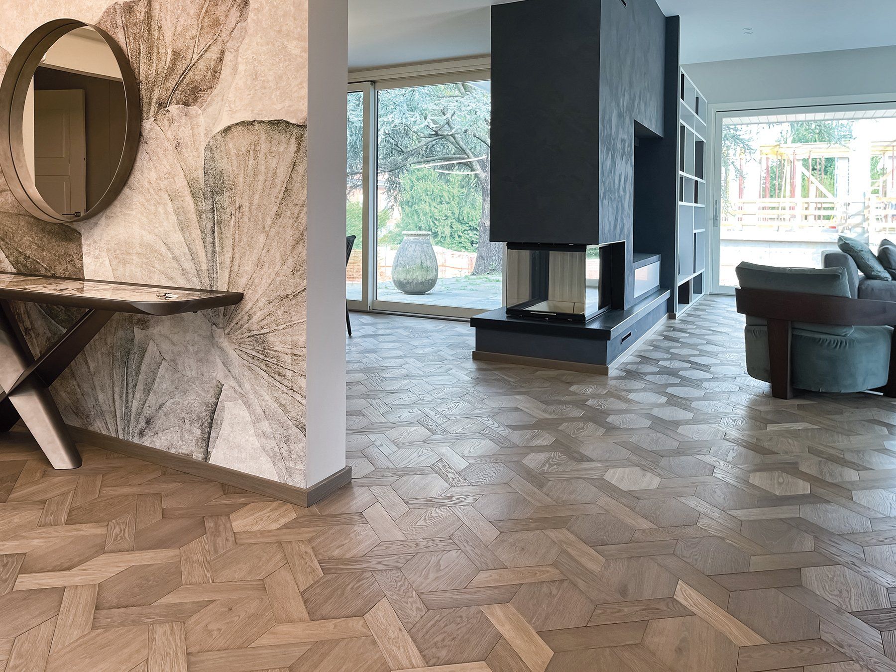 Ingresso con pavimento in legno decorato, carta da parati a motivi, consolle scura con specchio e camino.