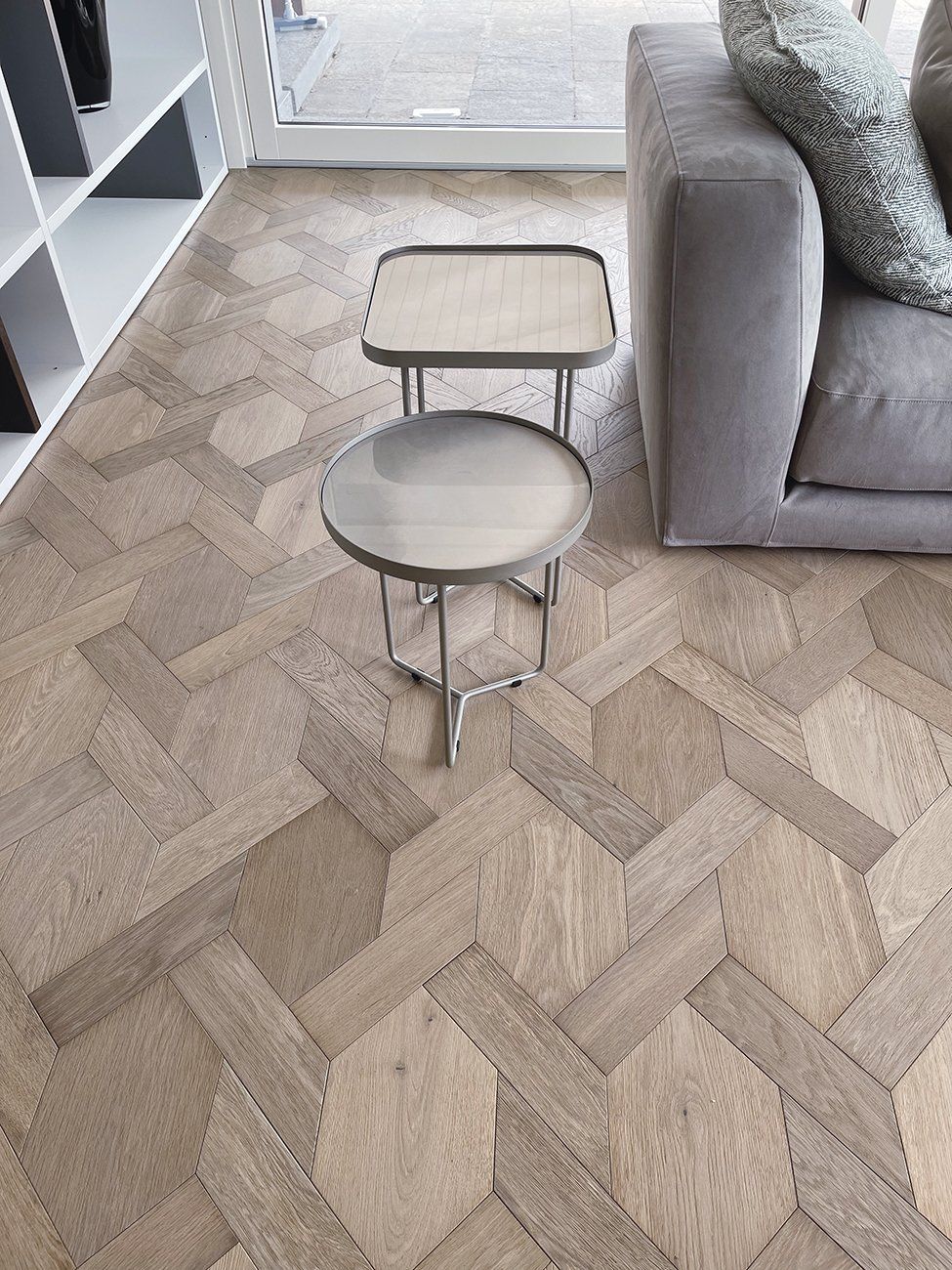 Pavimento in parquet di legno chiaro con due tavolini con gambe argentate vicino a un divano grigio.