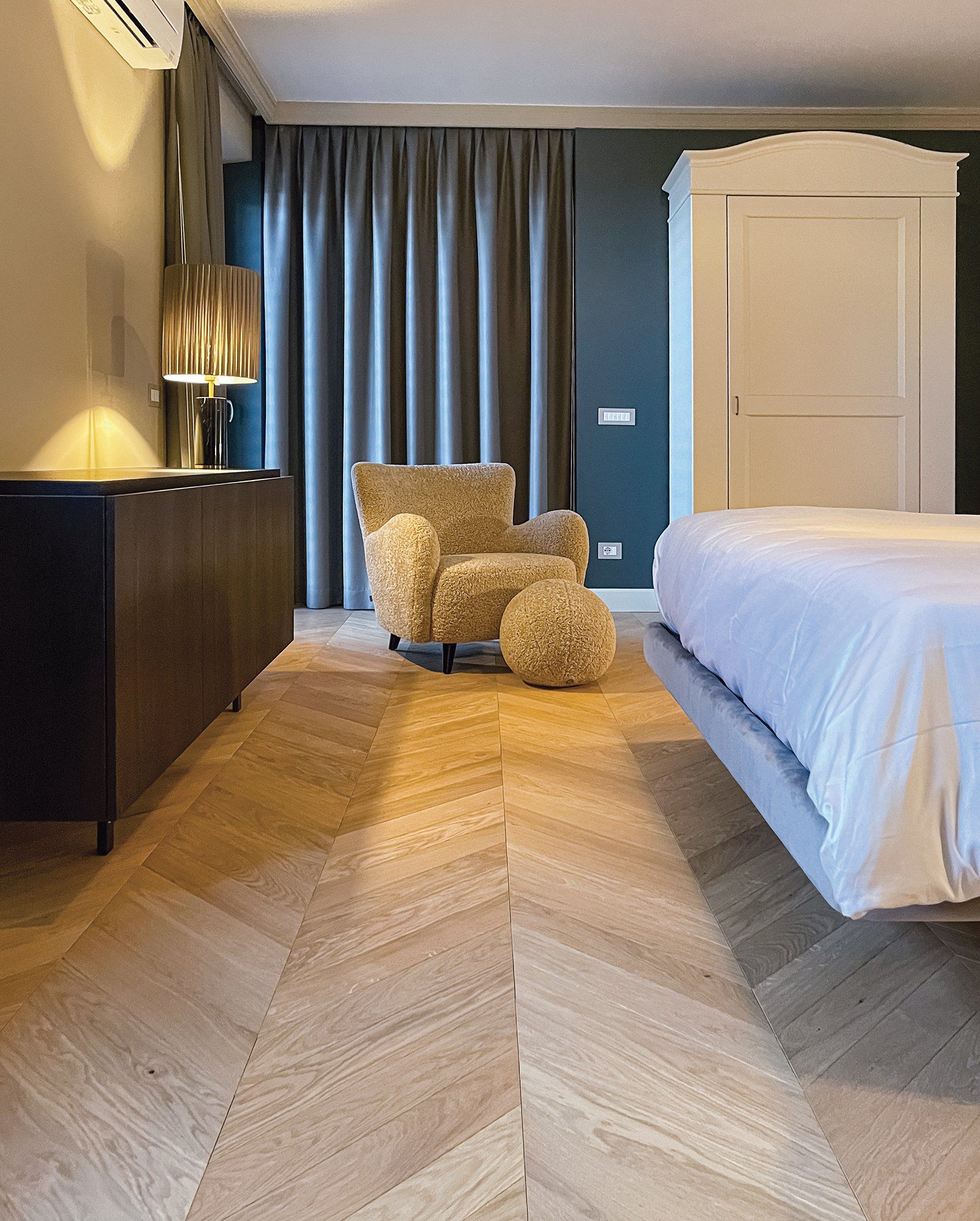 Camera da letto con pavimento in legno a spina di pesce, poltrona, tende scure e armadio bianco.
