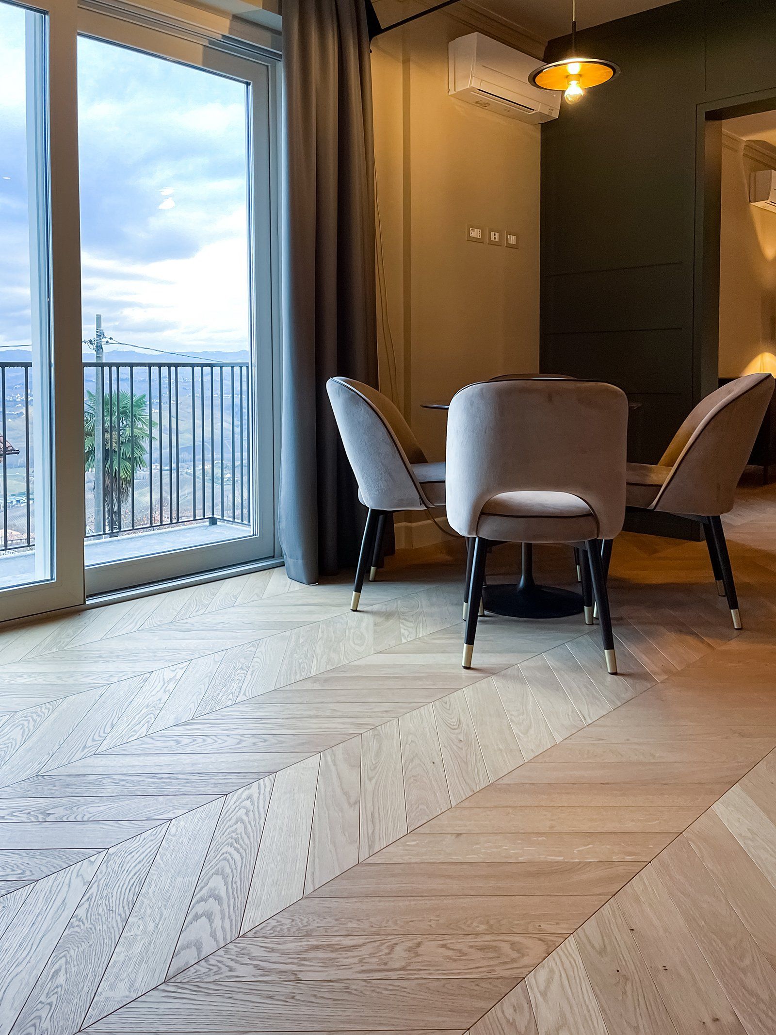 Sala da pranzo con tavolo, sedie e balcone. Pavimenti in legno chiaro, tende scure e cielo nuvoloso.