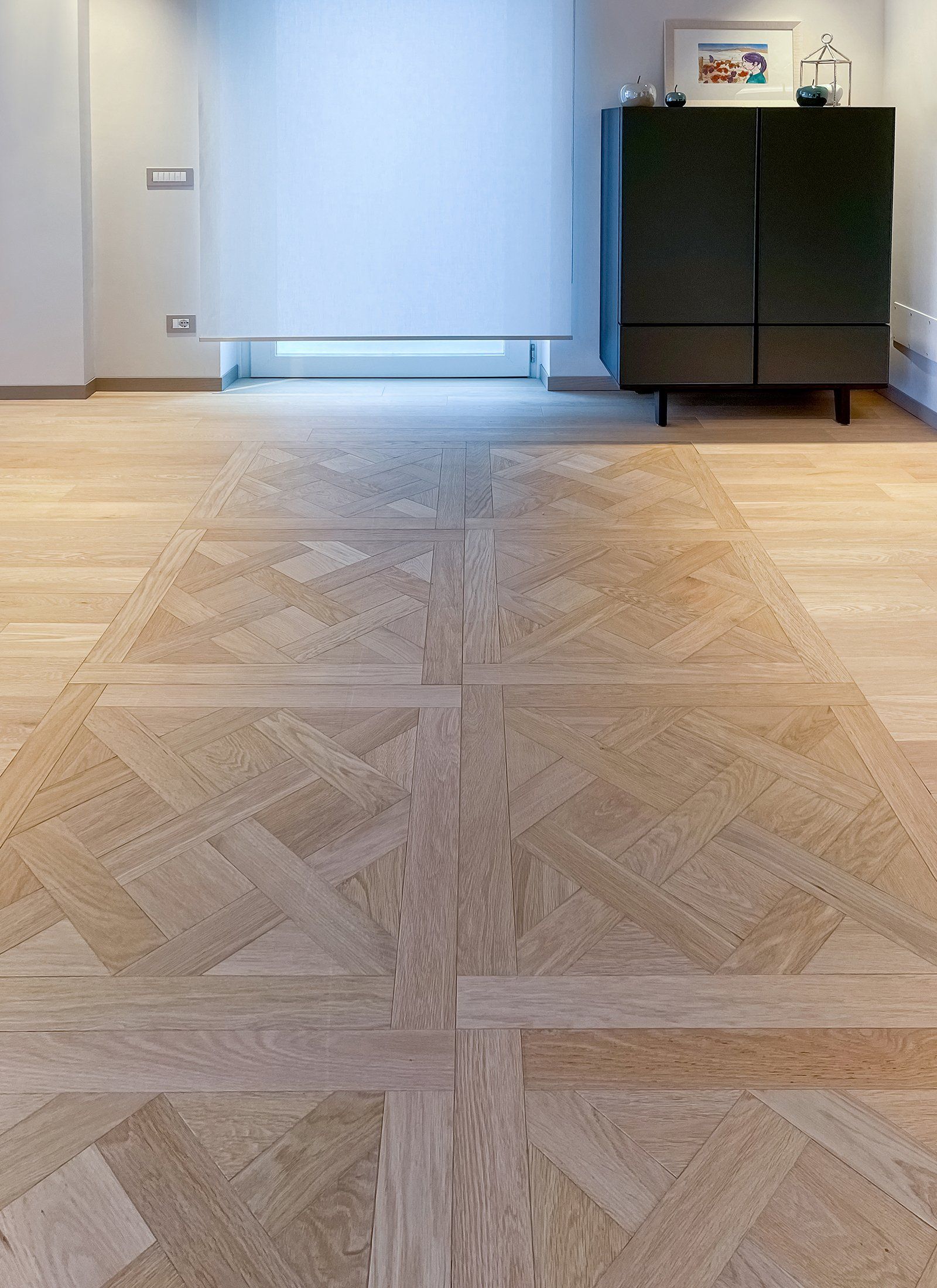 Pavimento in legno chiaro con motivo geometrico, parete bianca e mobiletto verde scuro.