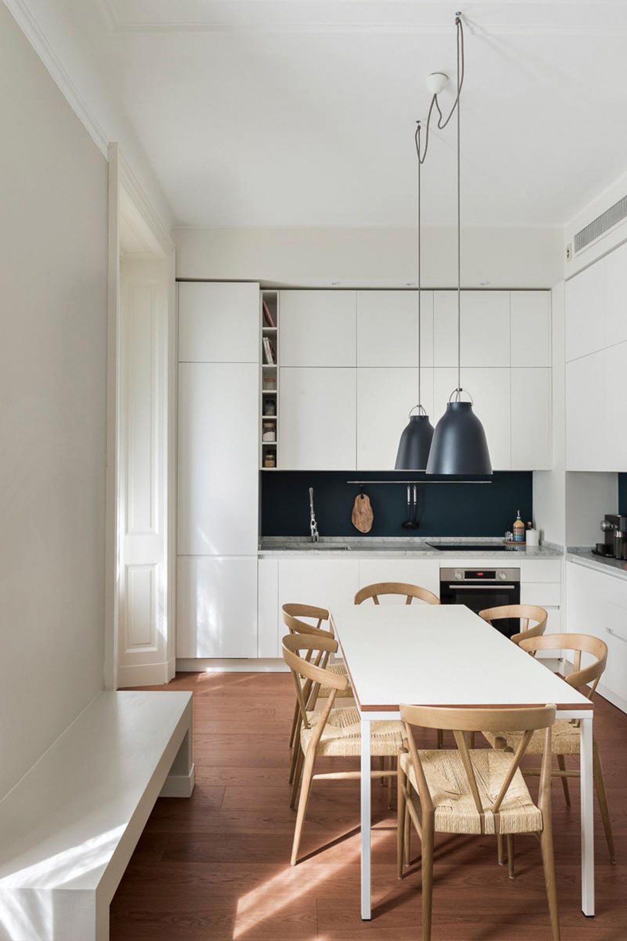 Moderna cucina bianca con lungo tavolo, sei sedie in legno e lampade a sospensione. Pavimento marrone-rossastro.