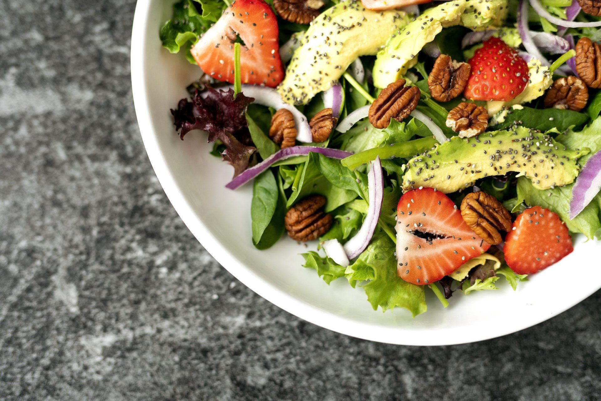 A salad with strawberries , avocado , pecans and onions on a white plate.