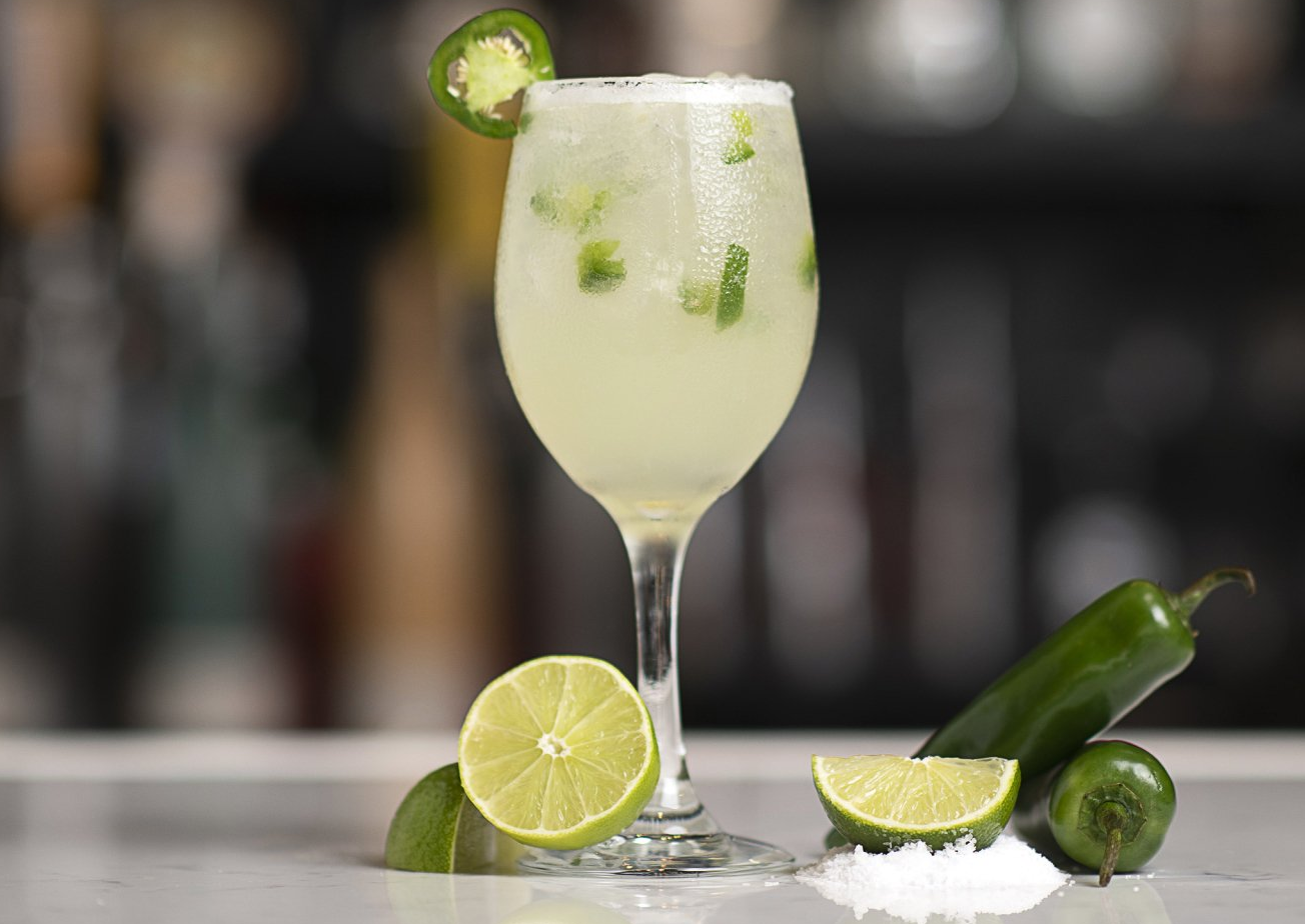 A margarita in a wine glass with limes and jalapenos on a table.