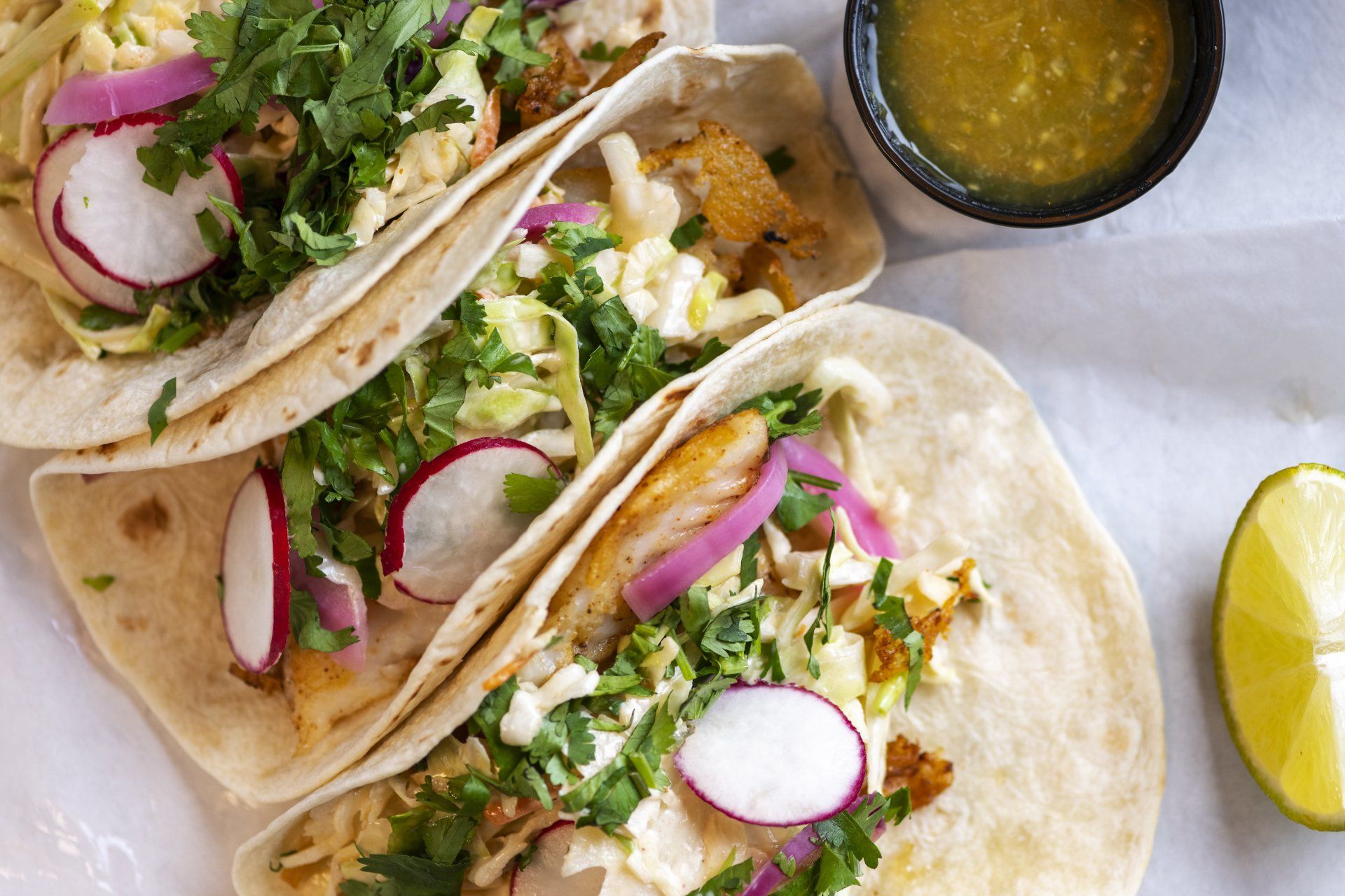 Three tacos are sitting on a table next to a bowl of salsa and a lime.