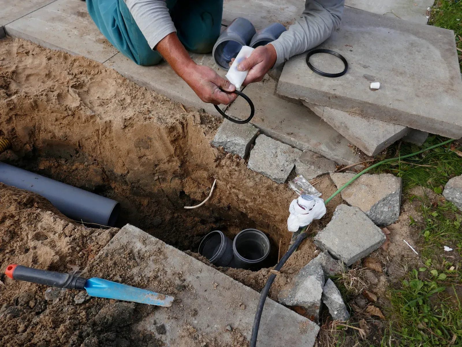 Hands installing underground wiring in a trench beside concrete slabs and PVC pipe connectors