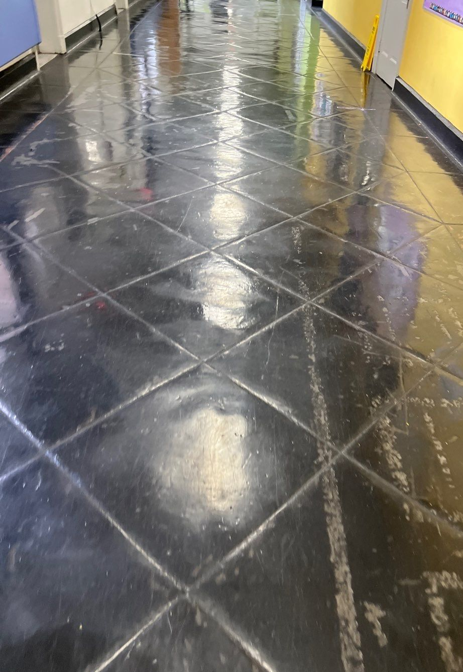 Shiny, black tile floor in a hallway with diagonal pattern. Reflections of lights and surrounding walls.