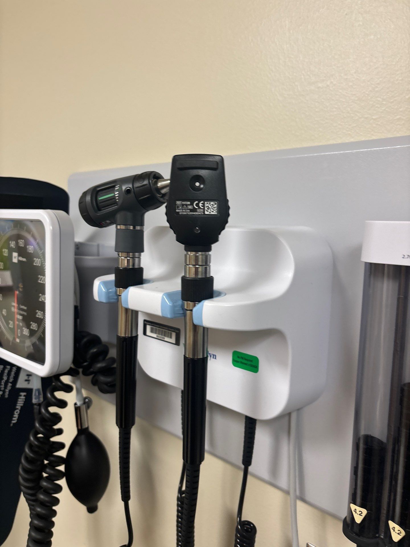 Medical examination tools in a wall-mounted holder: otoscope and ophthalmoscope. White, gray, black, and beige.