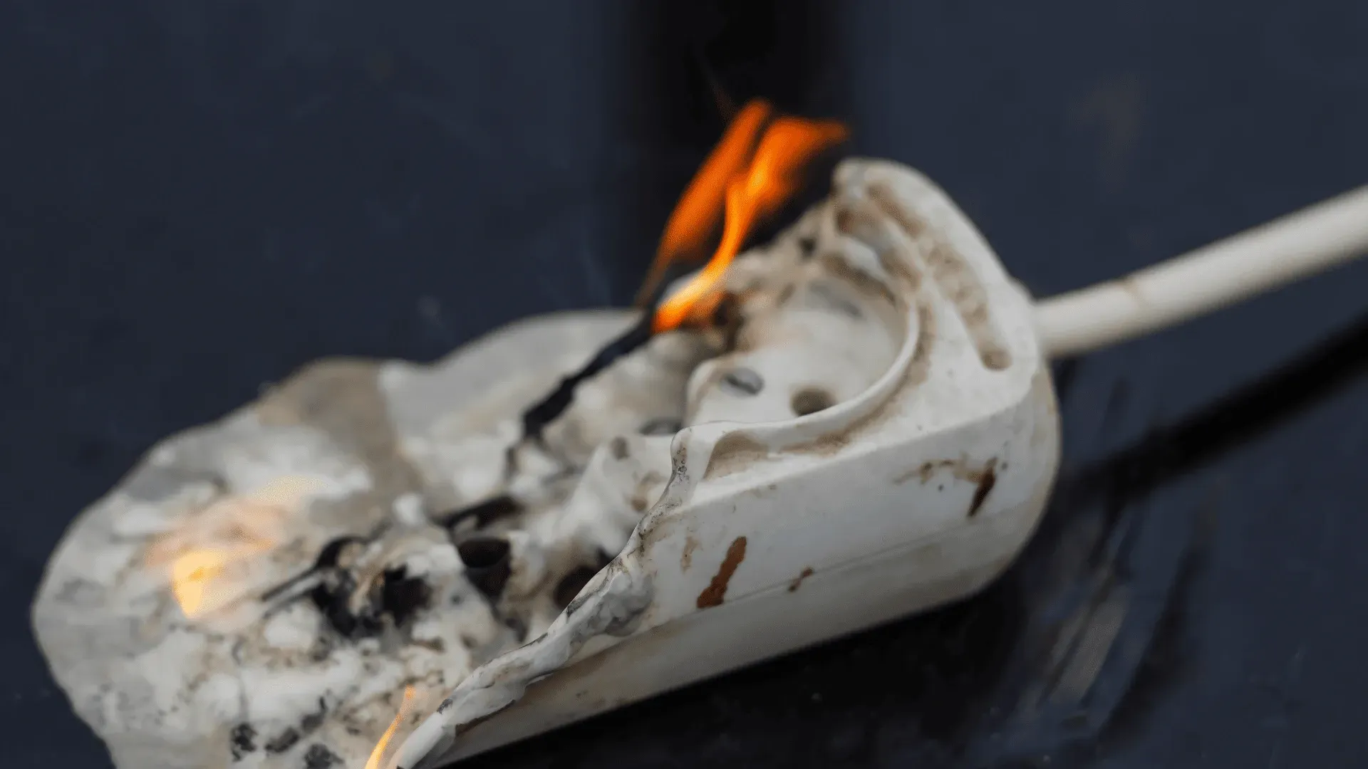 White plastic scoop on fire against a dark background, flames and smoke visible.