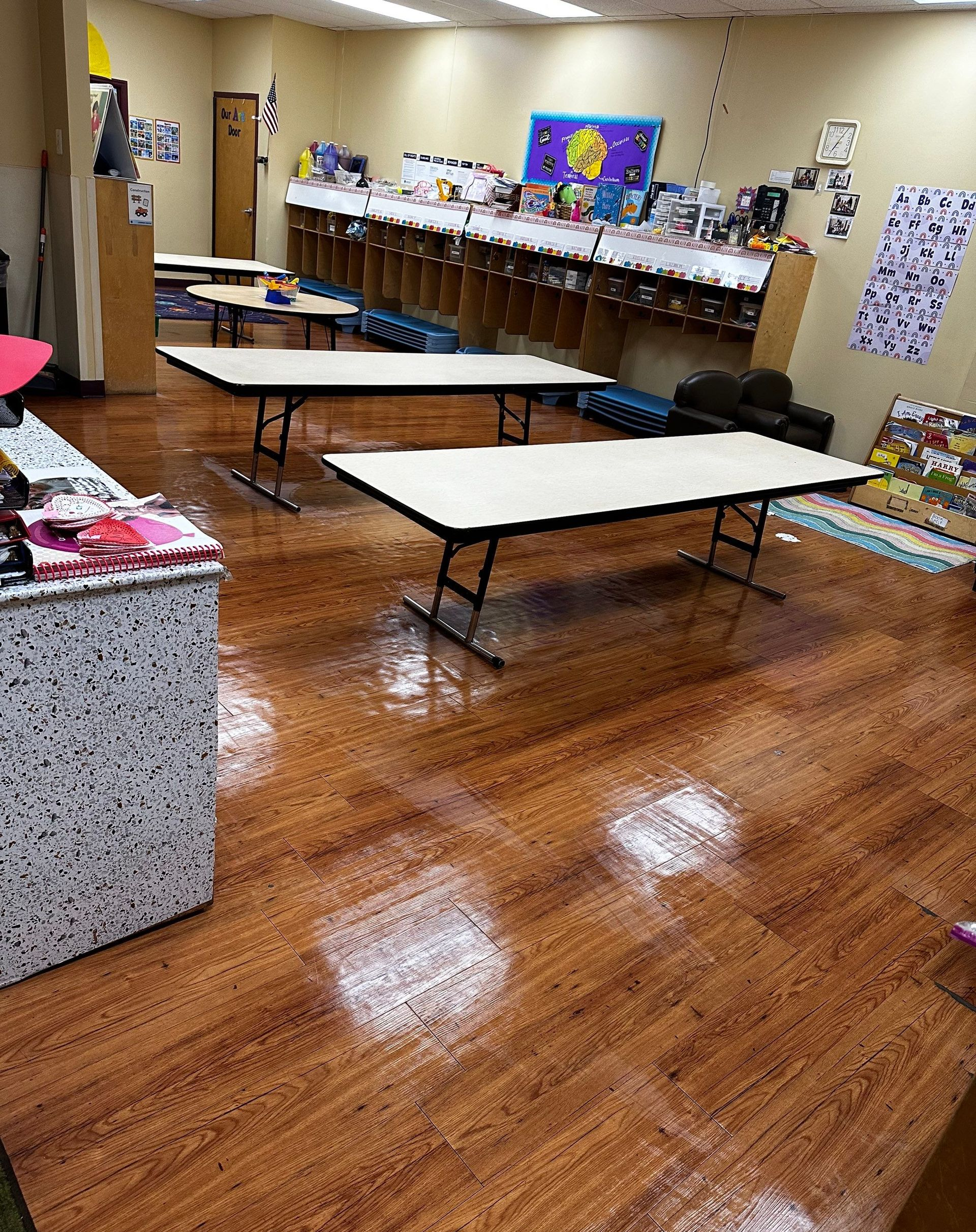 A classroom with wood-look flooring, tables, and shelves filled with supplies.