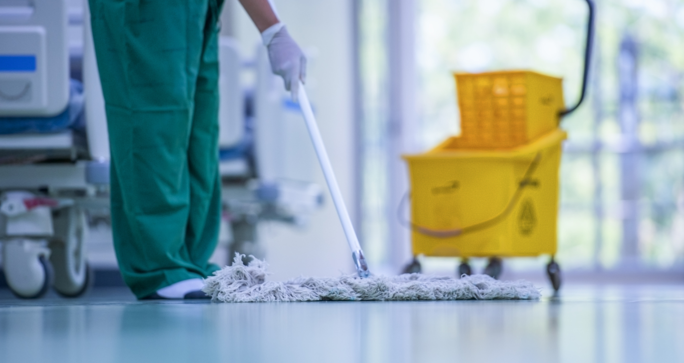 Person in green scrubs mops a hospital floor near a yellow cleaning cart.