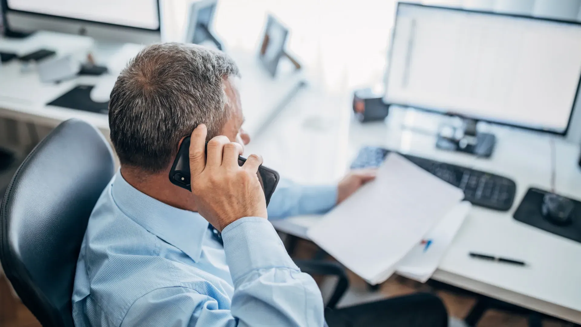 Man in blue shirt on phone, looking at papers in a home office with computer and desk.