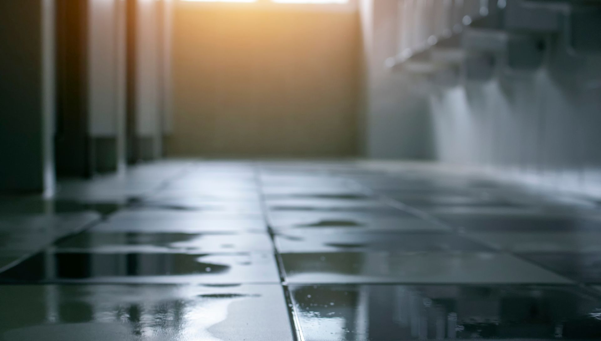 Wet tiled floor in a public restroom, with light shining from the end of the corridor.