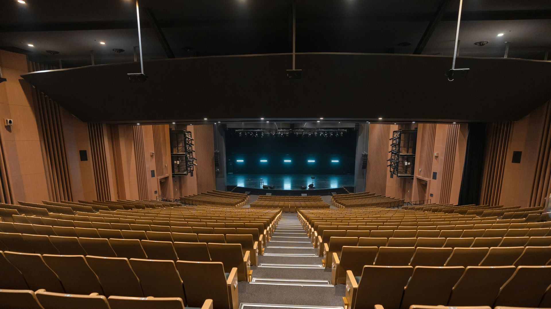 View from the back of a large, empty theater showing rows of tan seats facing a dimly lit stage with blue spotlights.