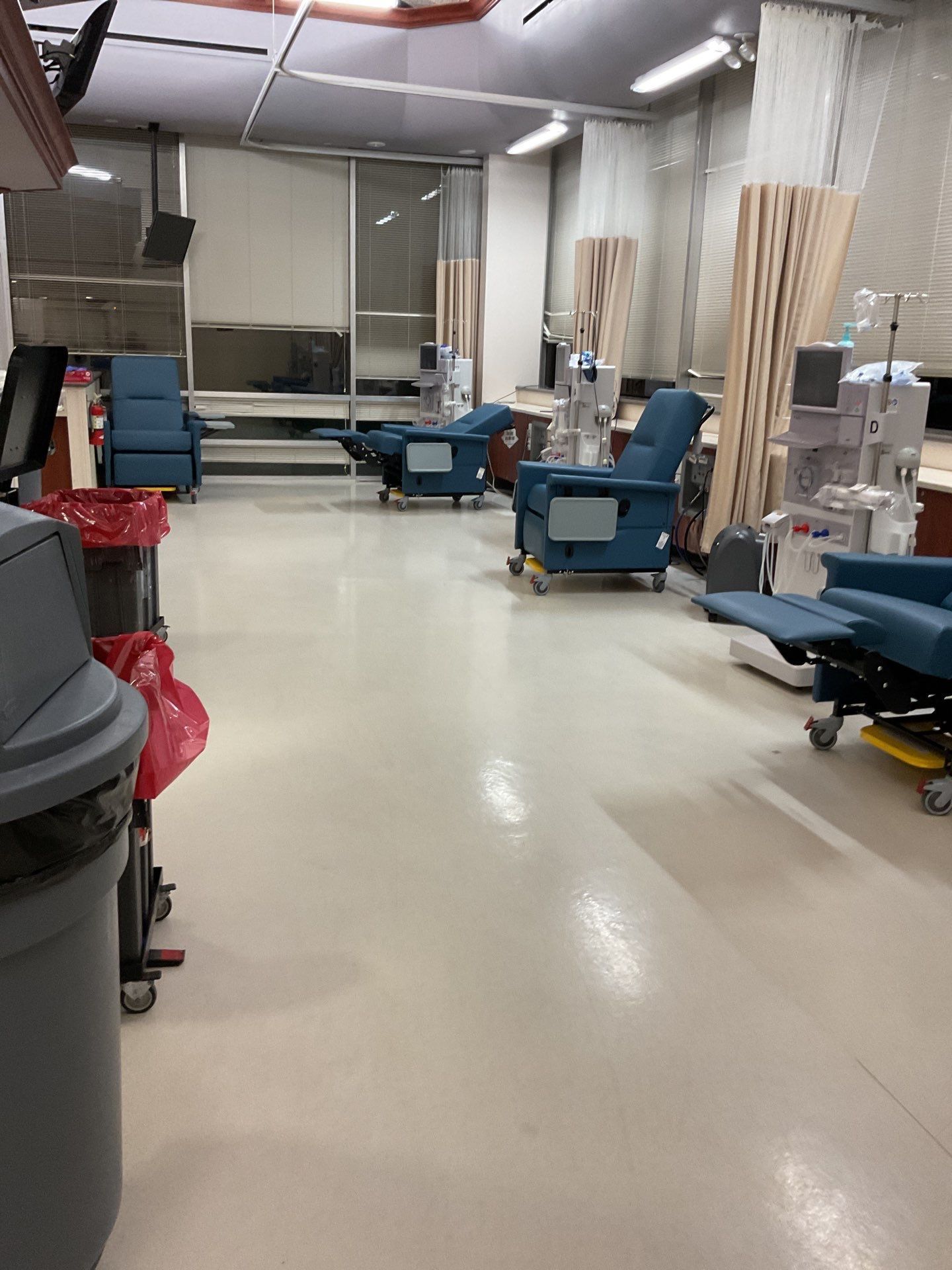 A dialysis room with blue chairs, medical equipment, and a window, illuminated by overhead lights.