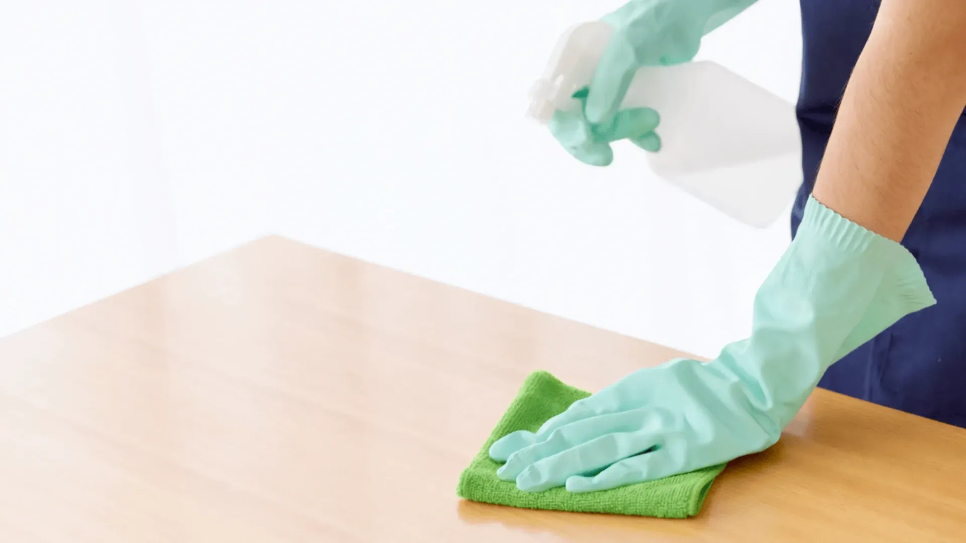 Person wearing green gloves cleaning a light wood table with a green cloth and spray bottle.