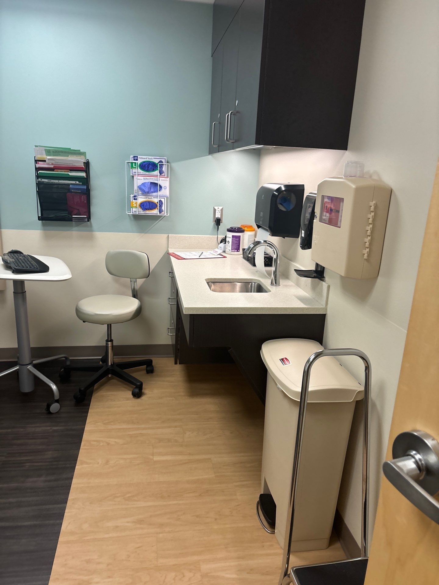 Examination room with sink, supplies, chair, and waste bin. Light blue and beige tones.
