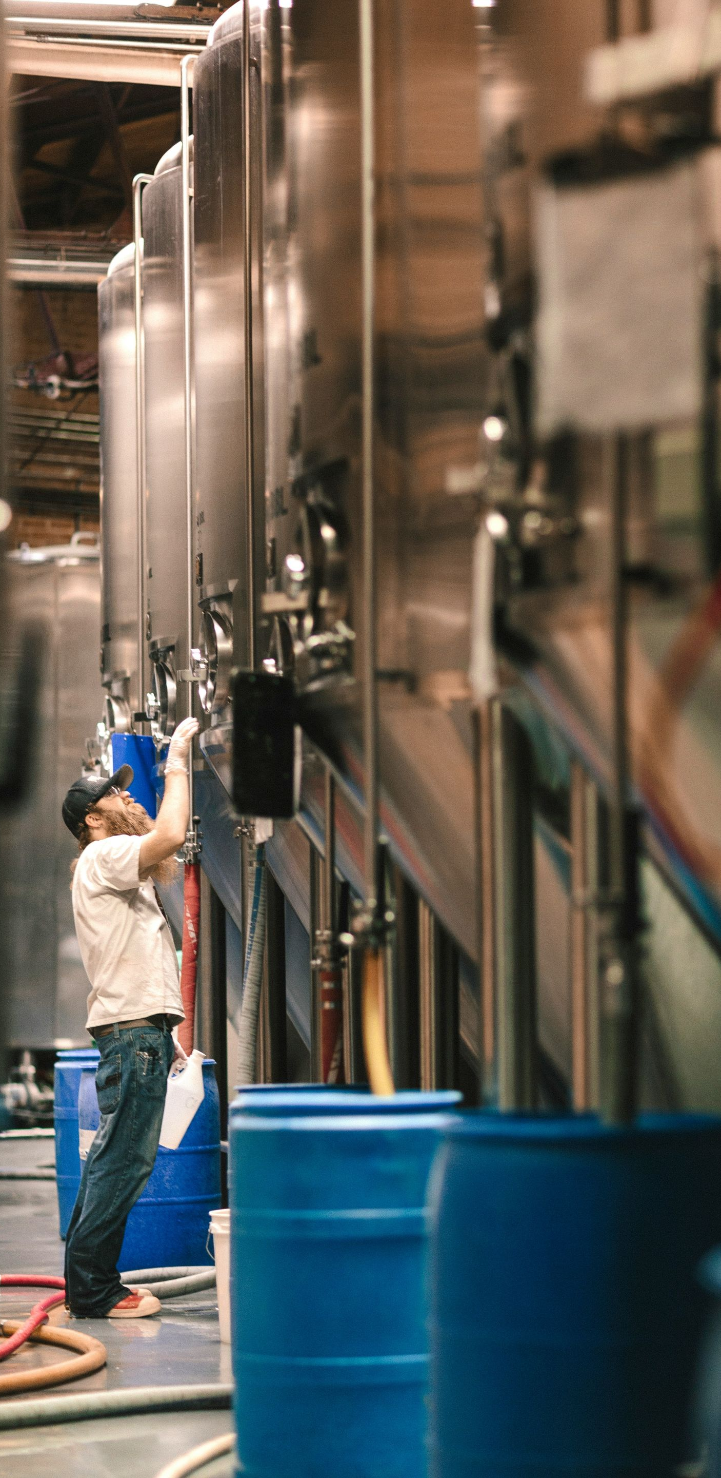 Person working on large metal tanks in a brewery, with blue barrels in the foreground.