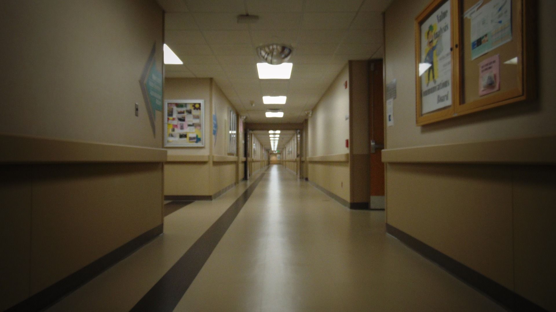 Empty hospital hallway with beige walls, fluorescent lights, and a dark floor stripe.