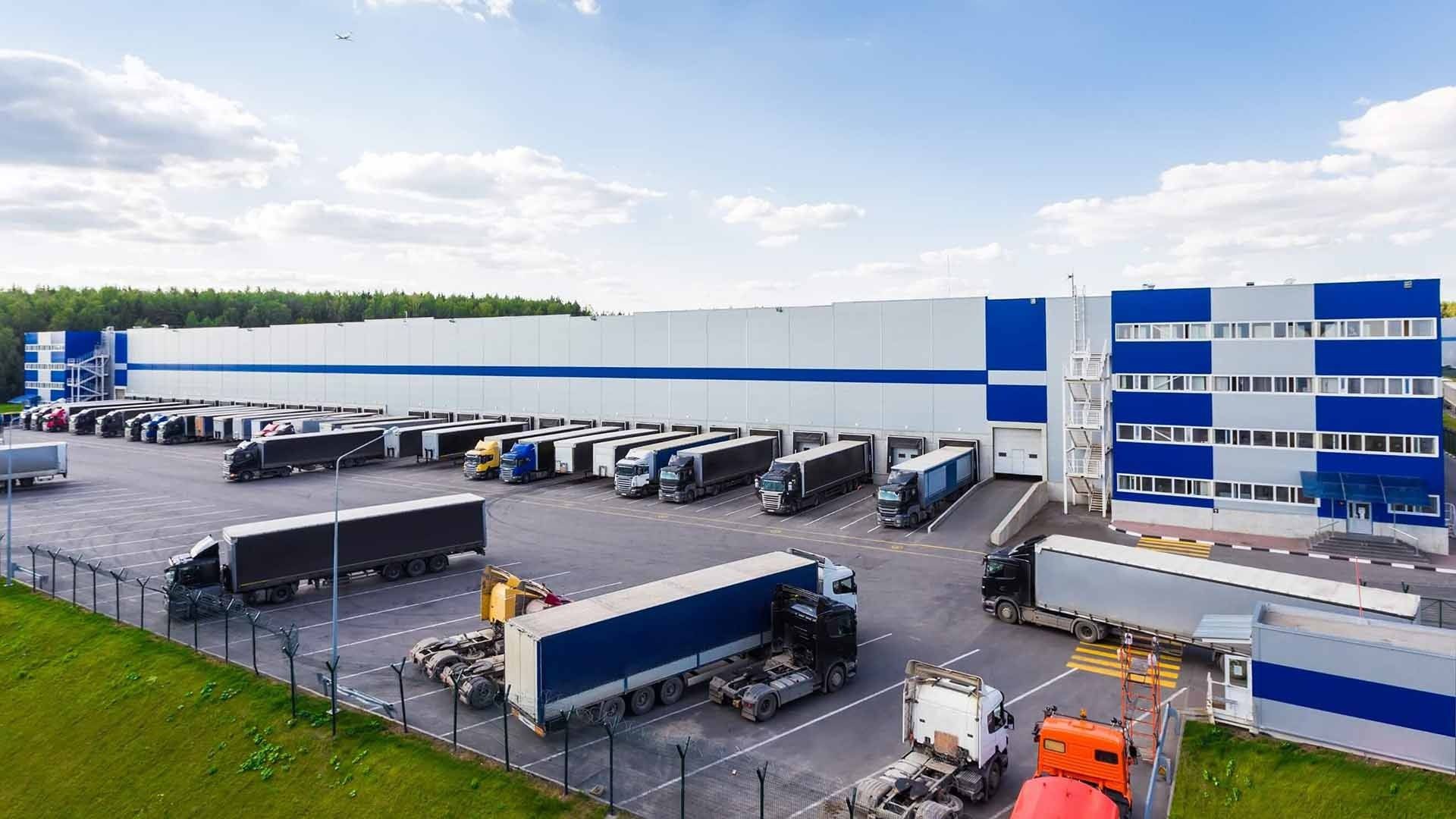 An aerial view of a large white and blue warehouse with numerous semi-trucks parked at loading docks in a paved lot.