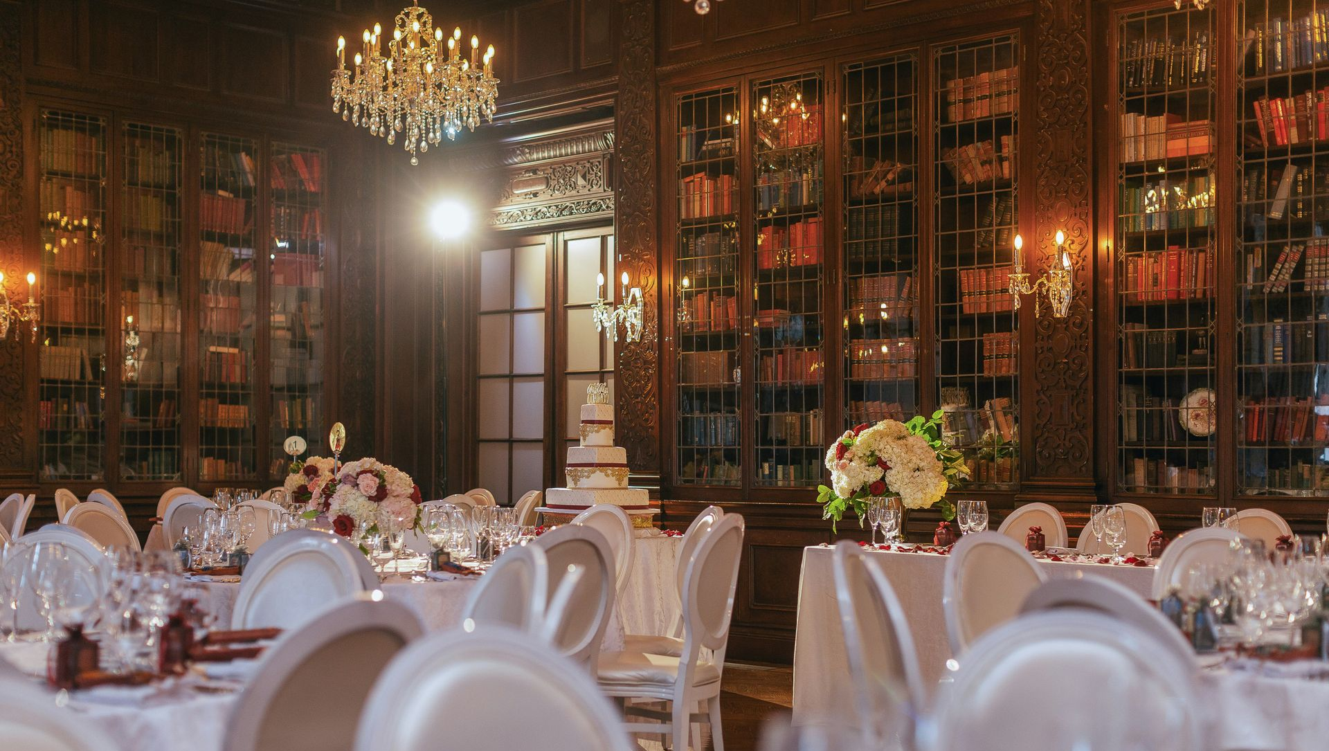 A dimly lit, wood-paneled library set for a wedding reception with round tables, white chairs, and floral centerpieces.