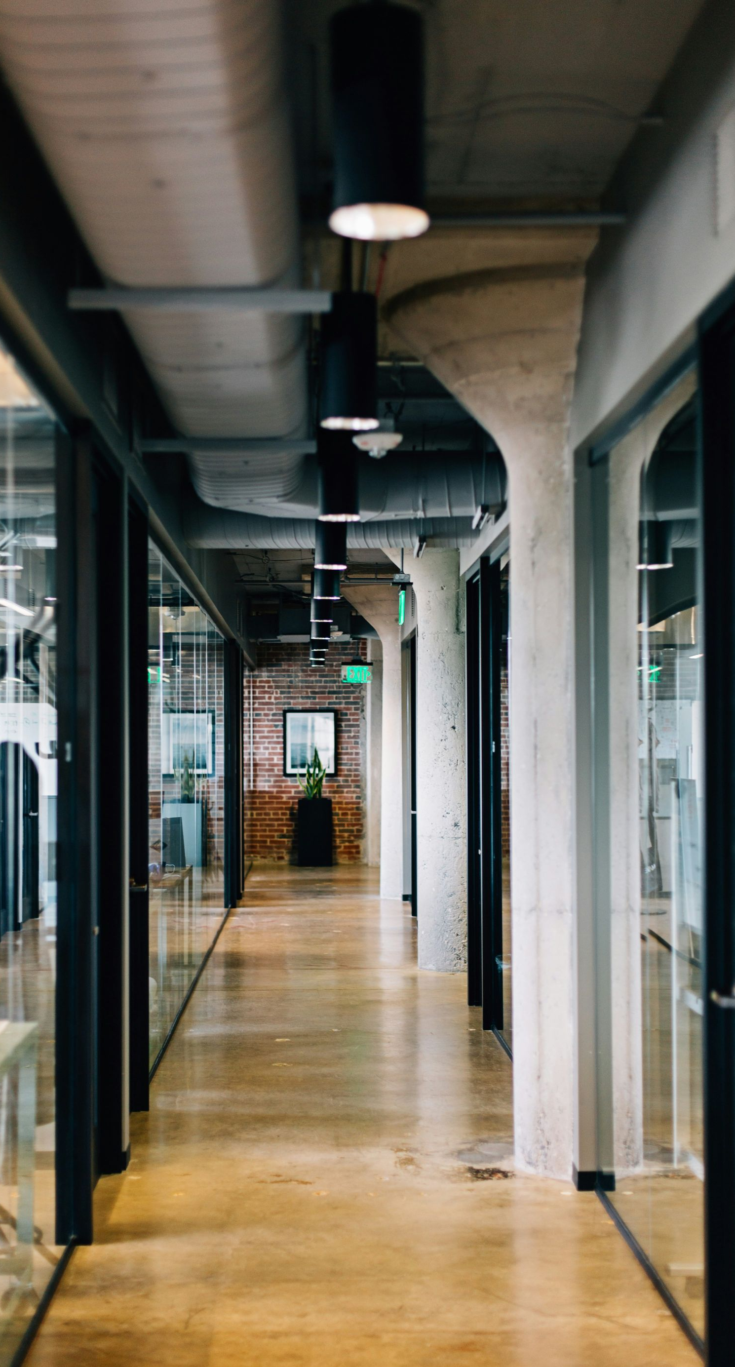 Office hallway with glass walls, wooden floor, and modern overhead lighting.