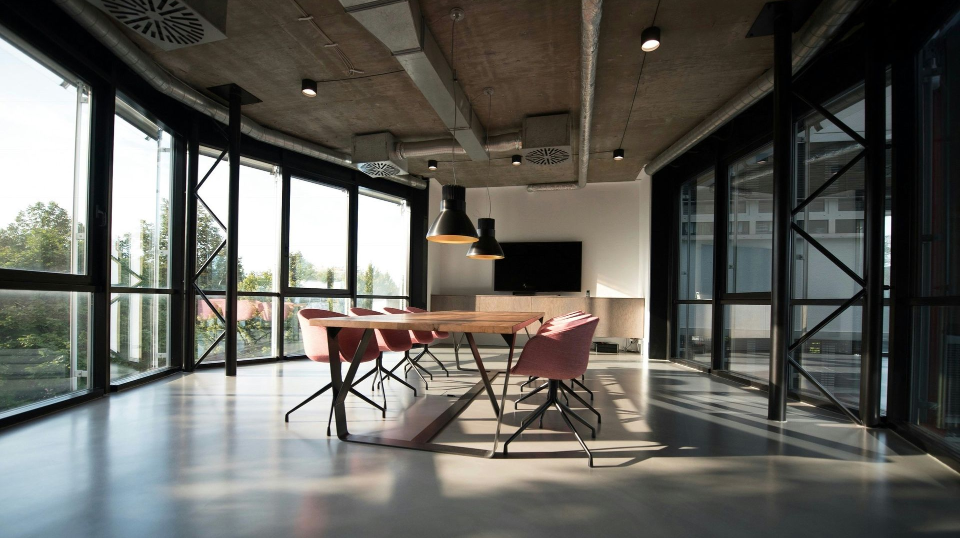 Modern meeting room with large windows, wood table, pink chairs, and exposed ceiling.