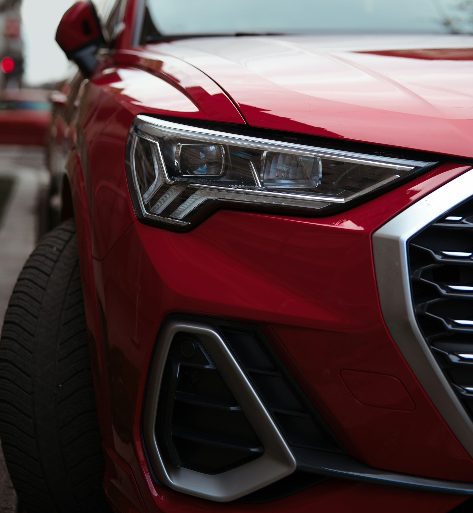 Red Audi Q3 car parked on a city street, close-up of headlight and grill.