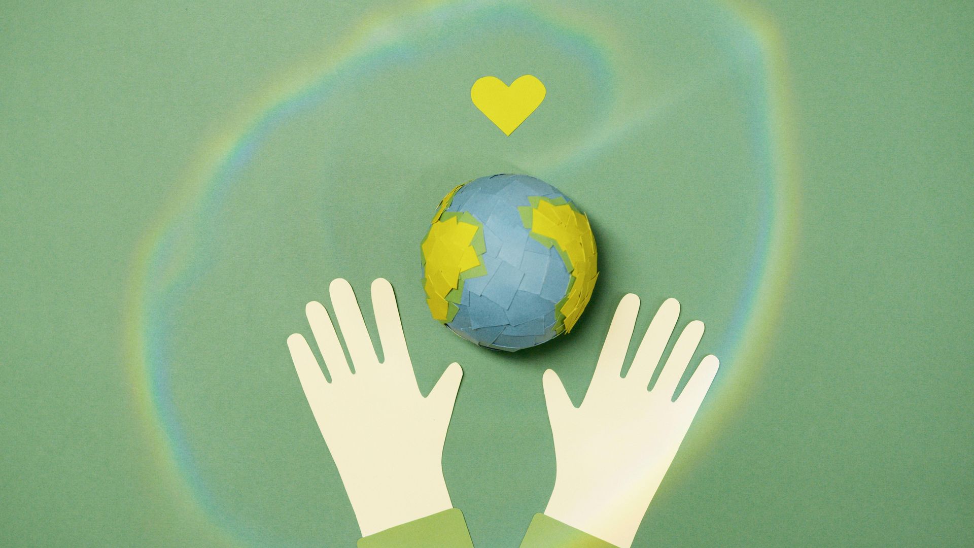 Hands reaching toward a paper-crafted Earth beneath a yellow heart, against a green background with a rainbow.