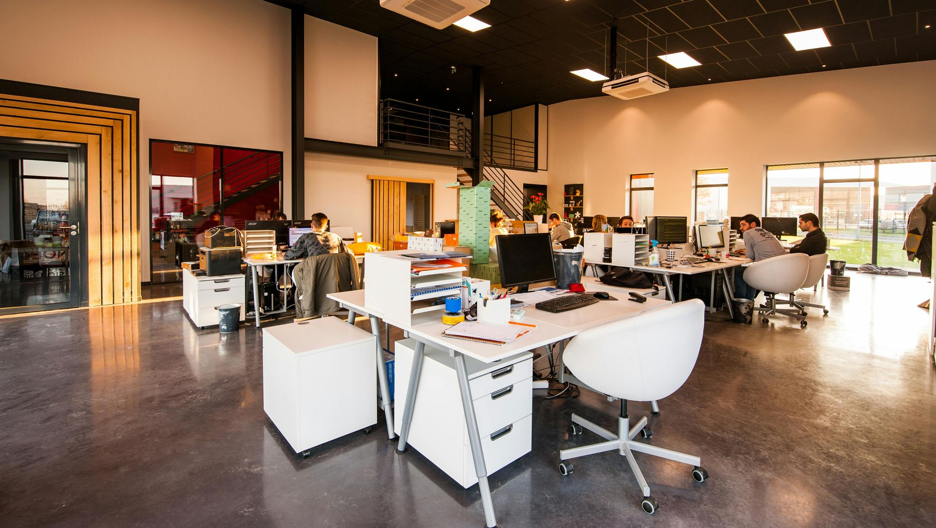 Open-plan office with people working at desks, under bright ceiling lights.